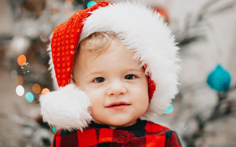 A baby wearing a santa hat in front of a Christmas tree enjoying Family Group Music Classes for Kids.