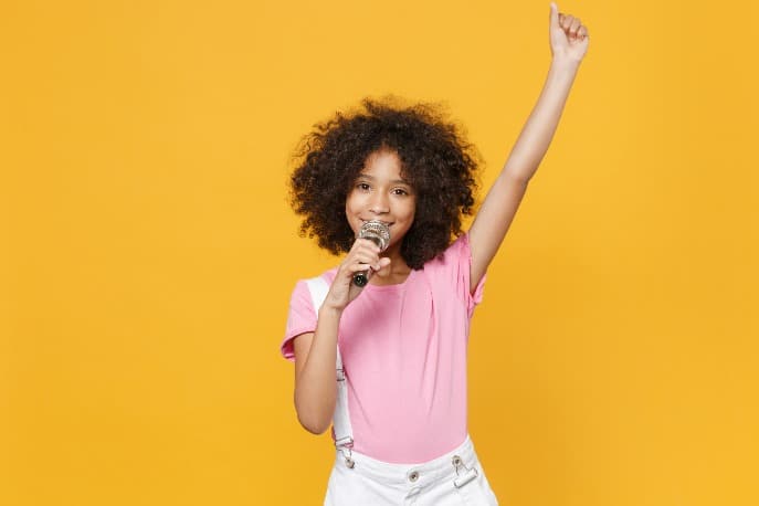 A young girl singing into a microphone against a yellow background.