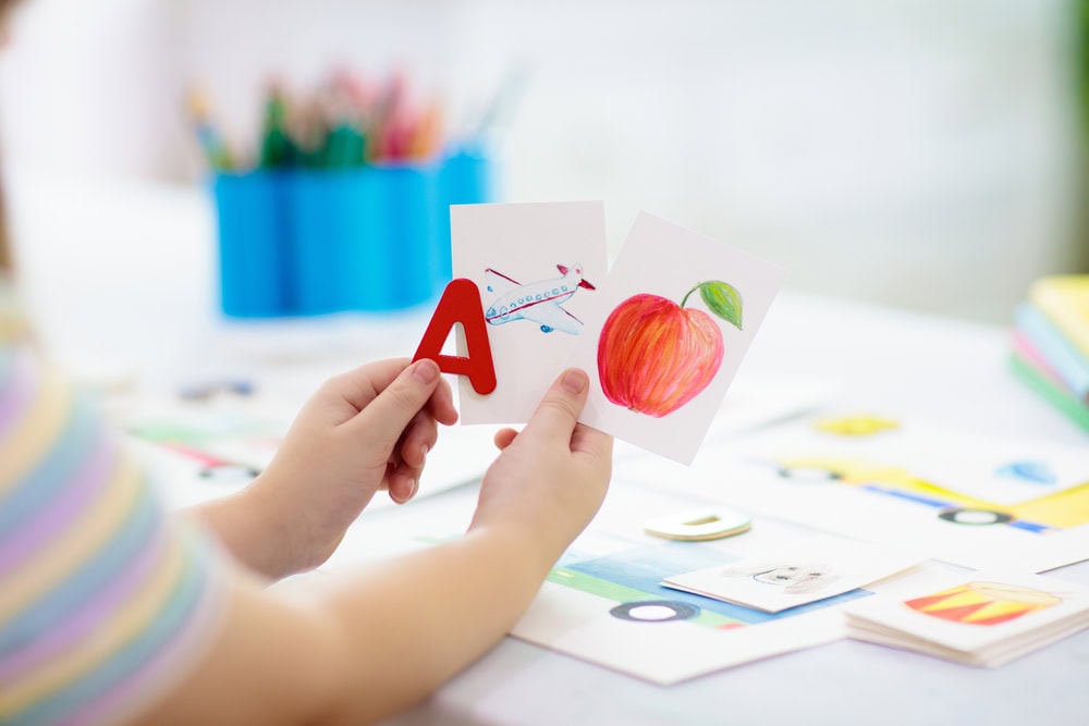 A child's hands over a table holding up the letter A and two drawings of an airplane and an apple