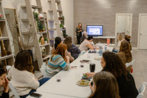 A group of women are seated at a long table watching another woman give a presentation in front of a monitor